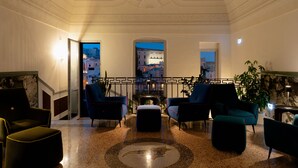 Een hal met een marmeren vloer, blauwe fauteuils, een centraal voetenbankje en uitzicht op een stadsgezicht door een boog.
