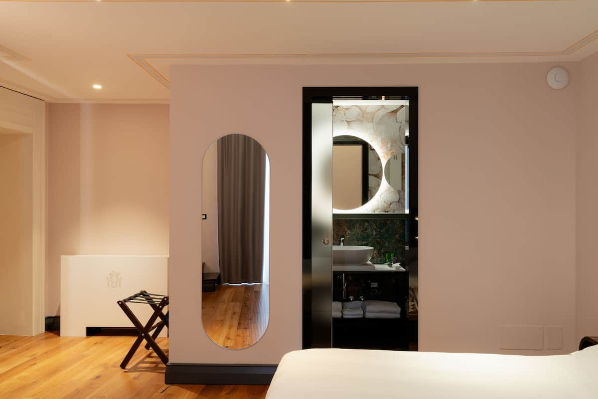 A modern bedroom with a large bed, a round mirror, and a built-in bathroom with a sink and towel rack.