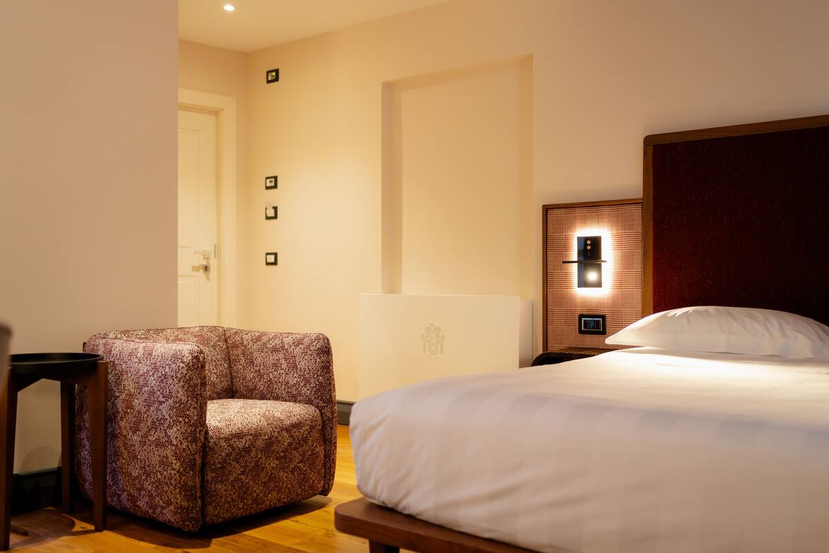 A hotel room with a bed, a chair, a desk, and a wall-mounted light fixture.