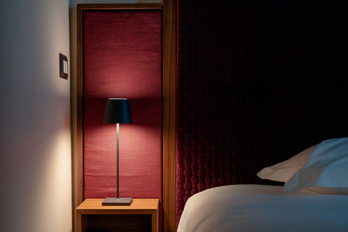 A hotel room with a bed, a nightstand, a lamp, and a red textured wall.