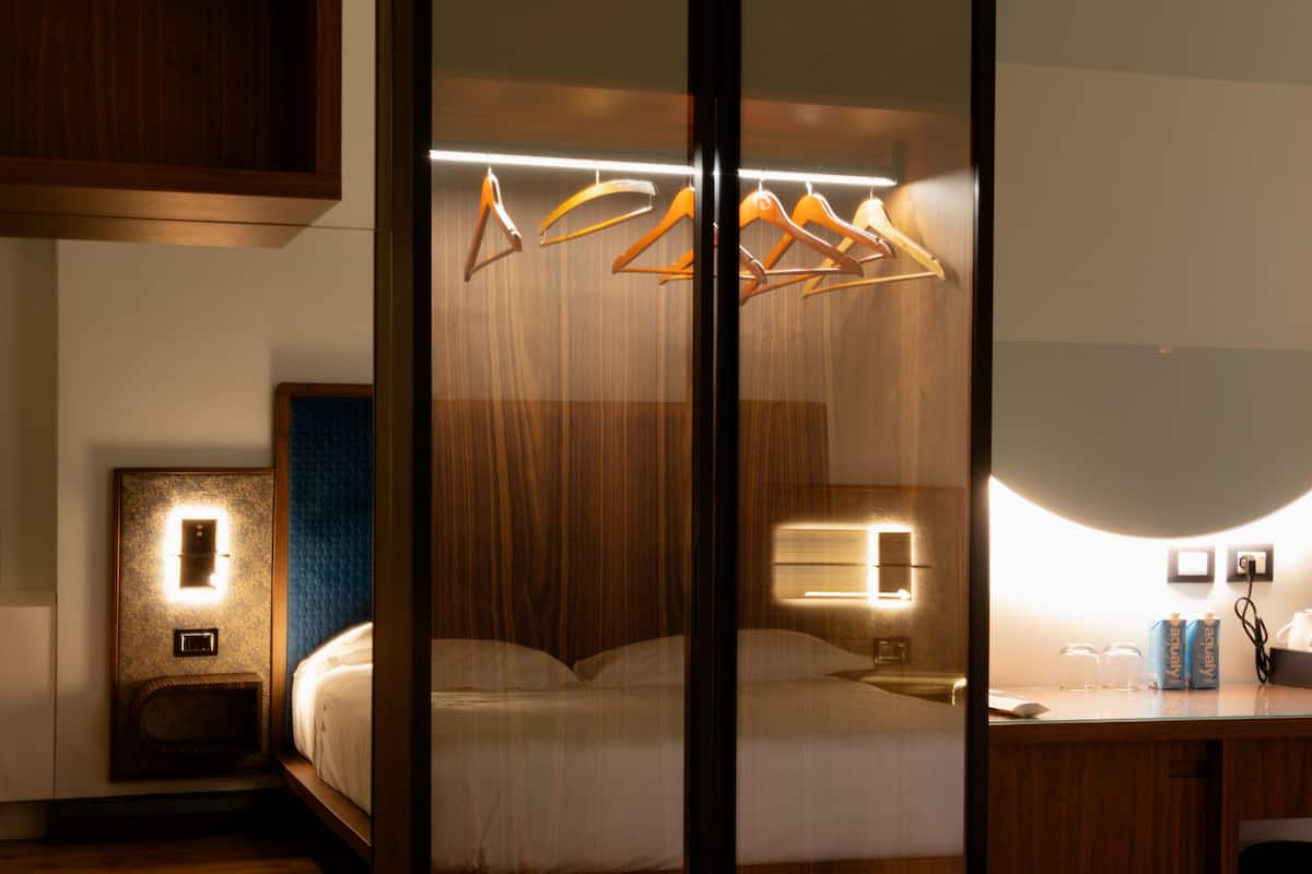 A modern hotel room with a large bed, a desk with a chair, a wardrobe, and a nightstand with a lamp.