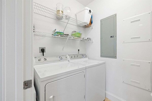 A laundry room with a front-loading washer and dryer, a shelf with cleaning supplies, and a small window.