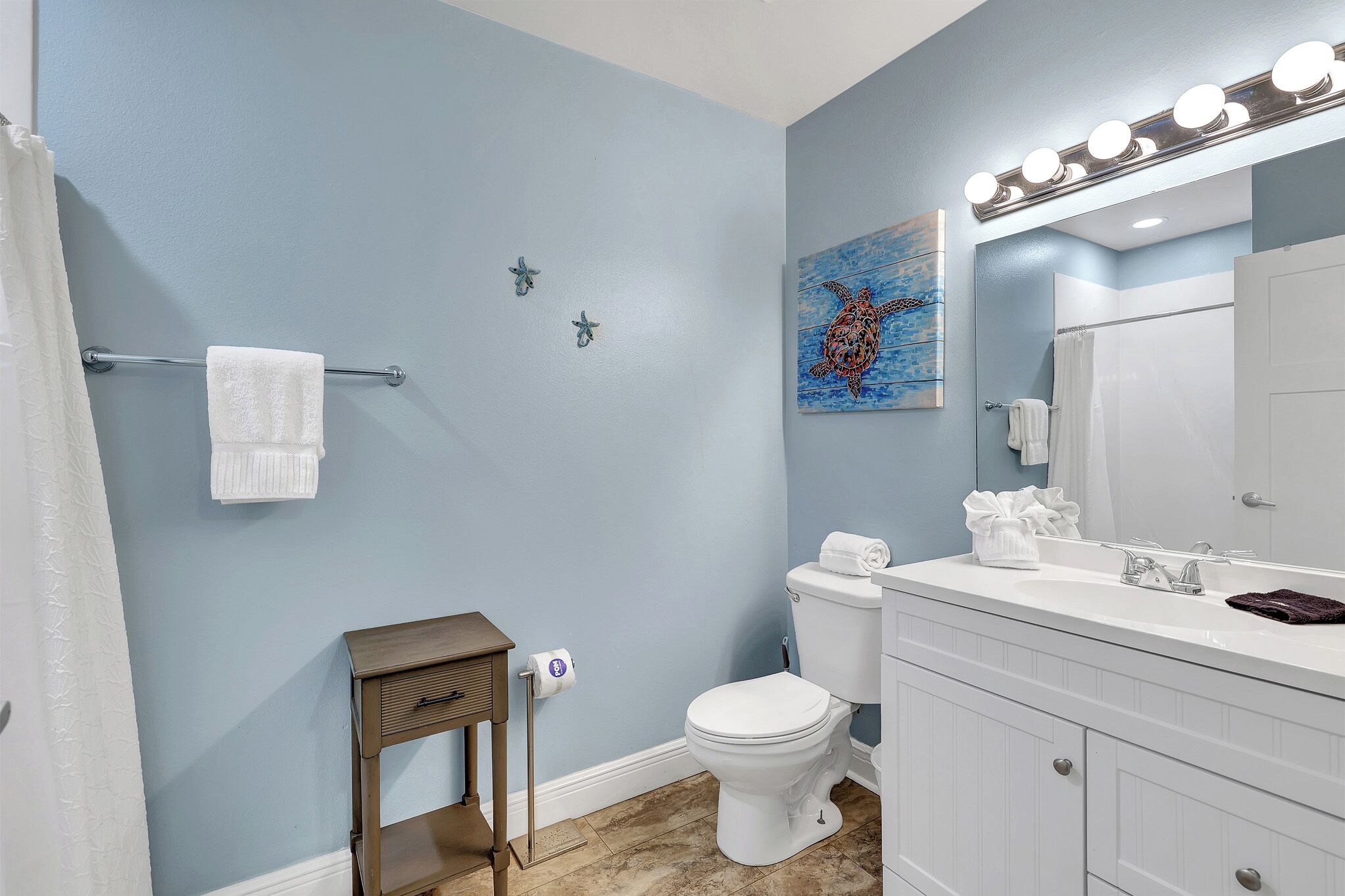 A bathroom with a toilet, sink, and vanity. There is a towel rack with a towel, a small table with a roll of toilet paper, and a painting on the wall.