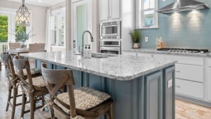 Une cuisine spacieuse dotée d’un grand îlot central, de meubles blancs, d’une hotte en acier inoxydable et de luminaires suspendus.