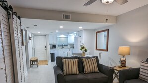 A living room with a sofa, a round ottoman, a ceiling fan, and a view into the kitchen. - NEW condo! Just 15 min to Ft Myers and Sanibel beach! Great Location!! (Fort Myers)