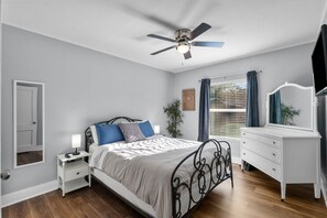 Un dormitorio con cama, mesita de noche, cómoda, ventilador de techo, ventana con cortinas y espejo.