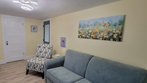 En stue med en grå sofa, en stilet lænestol, trægulv, en hvid dør og et blomsterbillede på væggen.