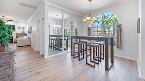 Ein modernes Wohnzimmer mit großem Fenster, einem Essbereich mit Holzhockern und einer gemütlichen Sitzecke mit Sofa und Topfpflanze.