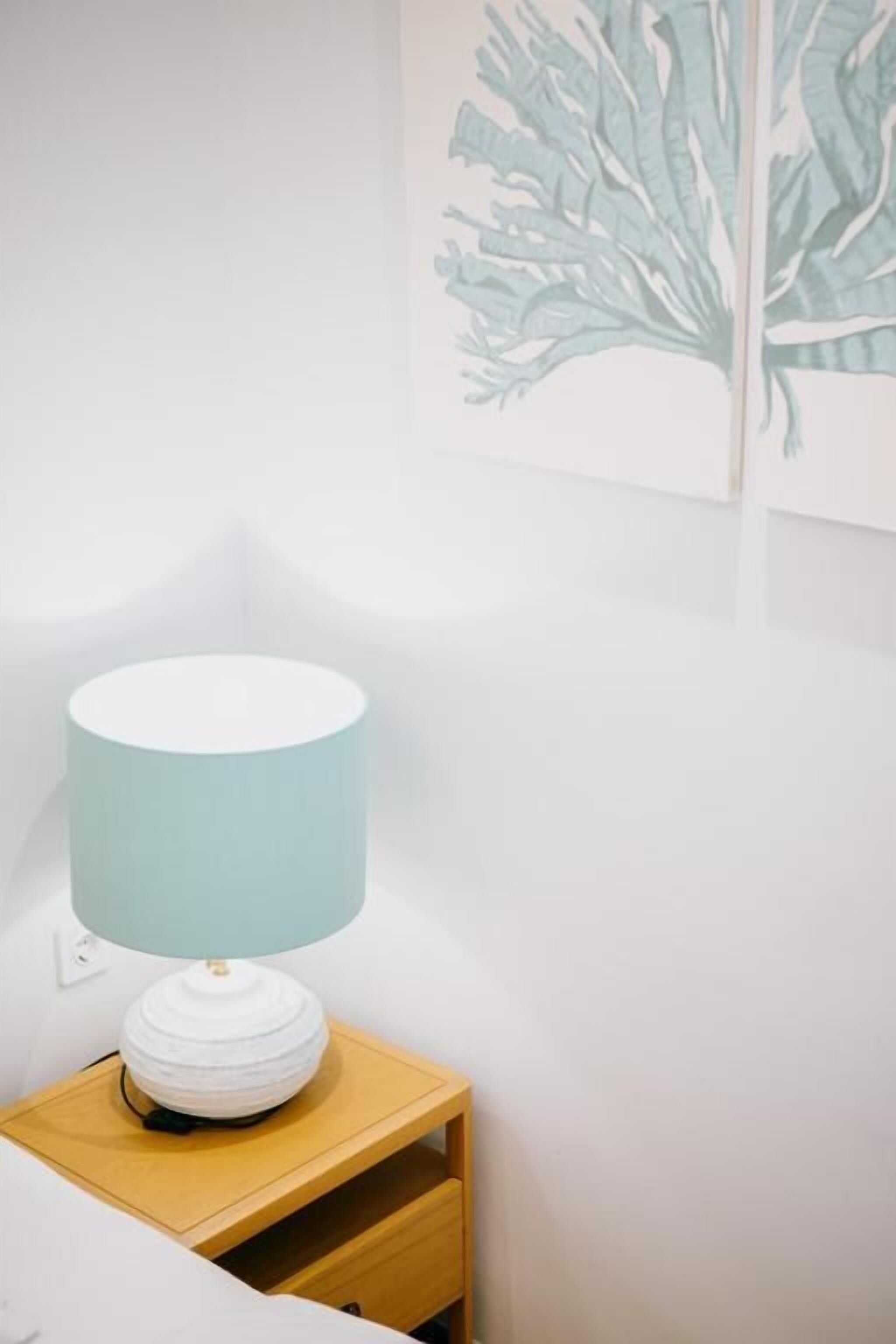 Een nachtkastje met een lamp, een muur met een bladmotief, en een witte muur.