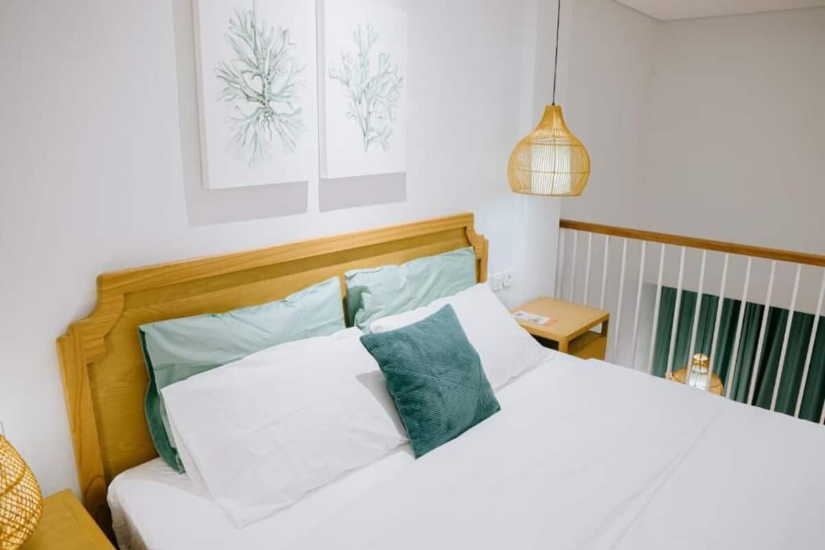 A neatly made bed with a wooden headboard, white and teal pillows, and a green accent pillow. There is a small bedside table with a lamp, and a hanging light fixture above the bed.