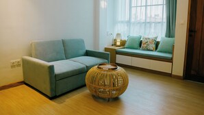 A modern living room with a grey sofa, a round wicker side table, and a window with sheer curtains. - Casa Amarta Canggu - Boutique Loft & Detox Spa (Canggu)