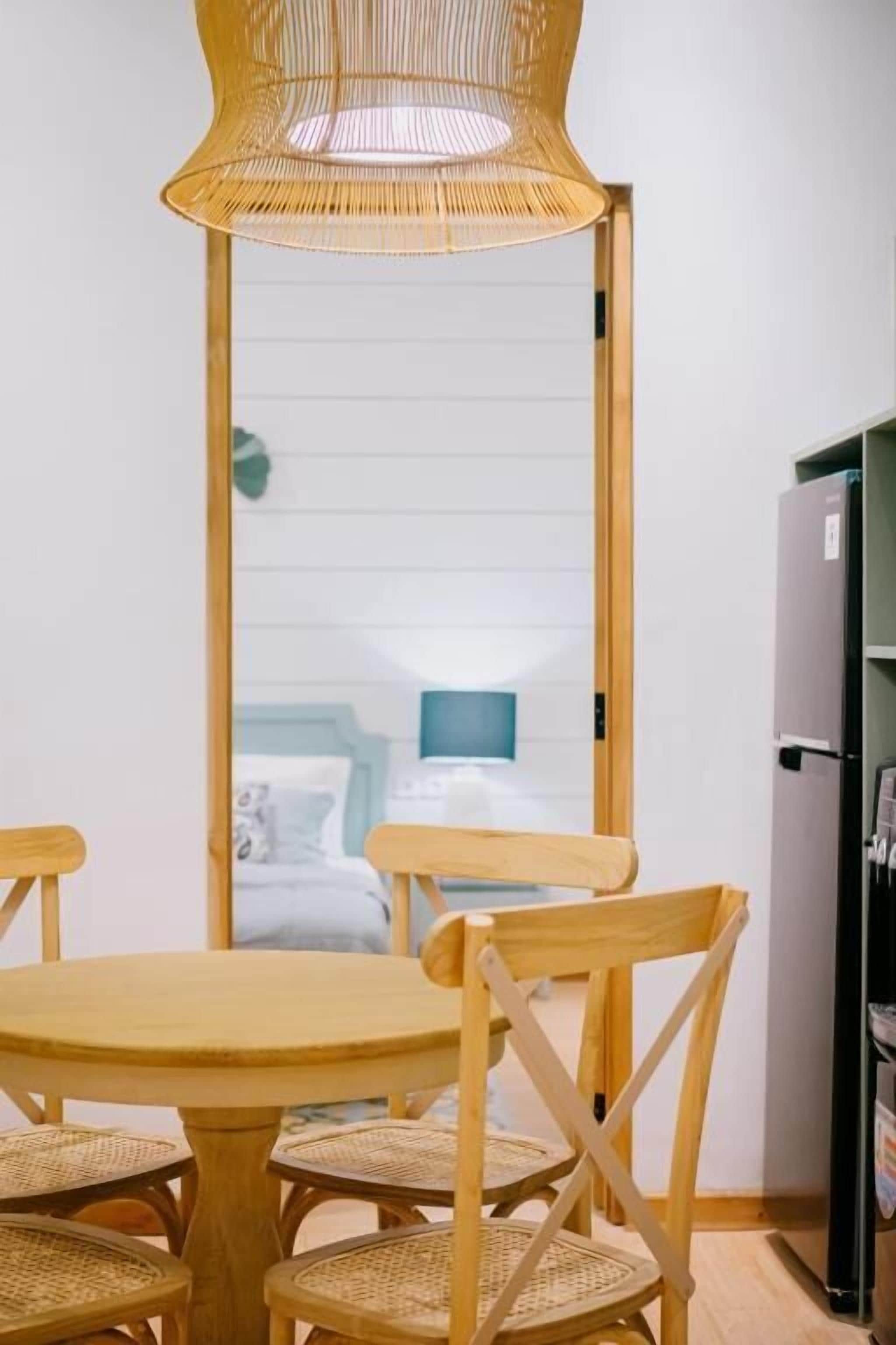 Een kamer met een ronde houten tafel en stoelen, een geweven hanglamp en een uitzicht op een bed via een spiegel.