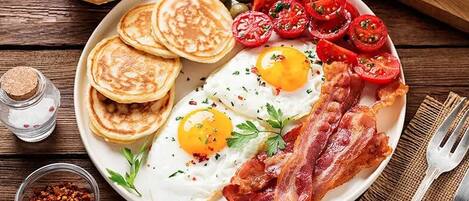 A breakfast plate with eggs, bacon, tomatoes, and pancakes on a wooden table.