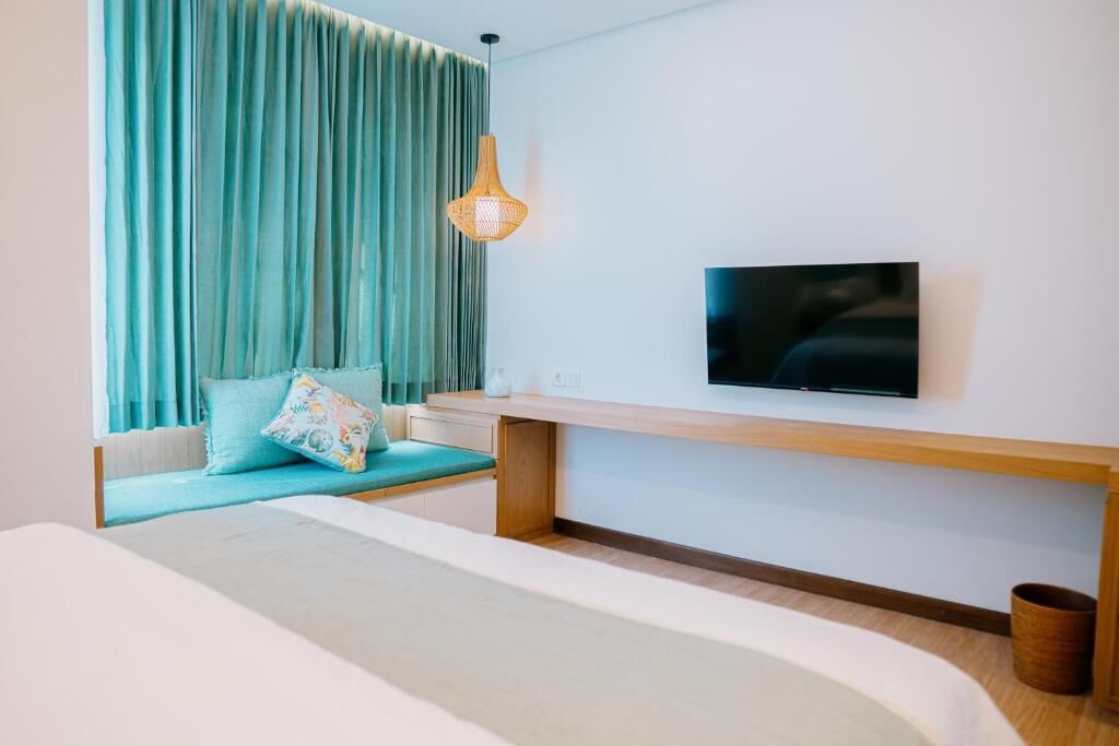 A modern hotel room with a large bed, a TV mounted on the wall, a wooden bench, and a window with curtains.
