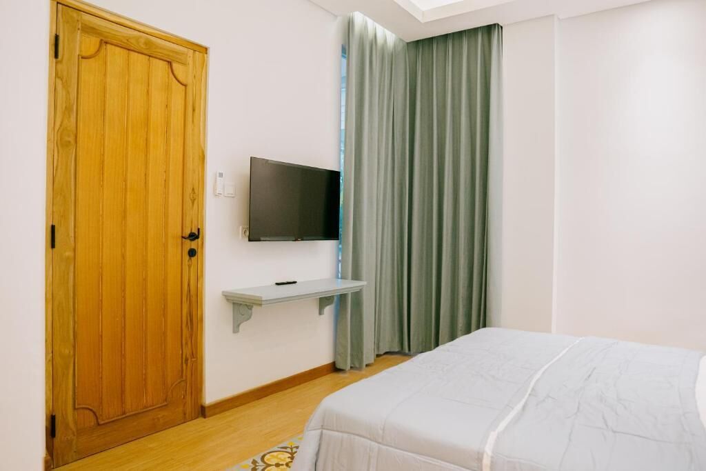 A bedroom with a wooden door, a mounted TV, a small shelf, a bed with a white bedspread, and a green curtain.
