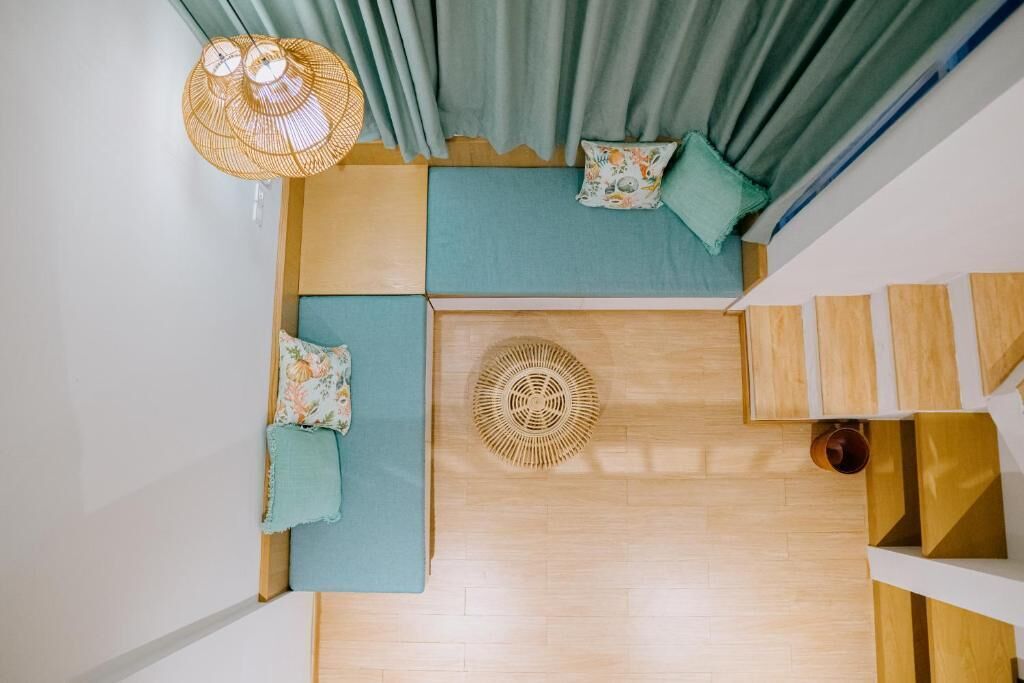 A top-down view of a room with a wooden floor, a woven floor lamp, a green sofa with cushions, and a small round table.