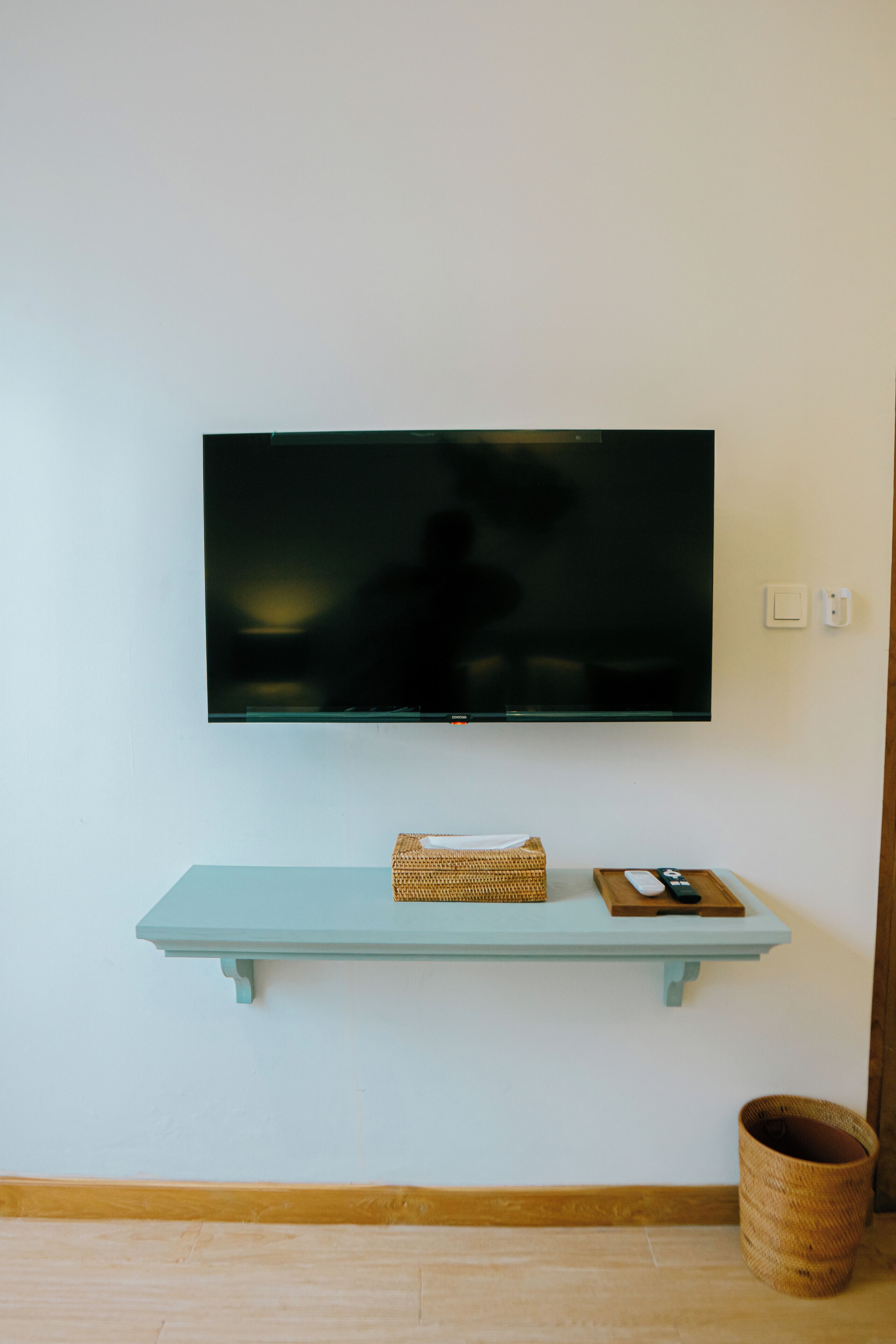 A flat-screen TV mounted on a wall above a light blue shelf with a basket and remote control.