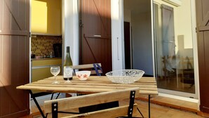Une porte coulissante ouverte donne sur un balcon où se trouvent une table en bois, deux verres à vin, une bouteille de vin et un bol décoratif.