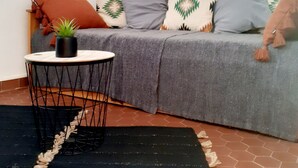 Un salon accueillant avec un canapé gris, des coussins à motifs, un miroir rond, une petite table ronde agrémentée d’une plante et un tapis noir.