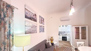 Un salon avec un canapé, une table et une lampe. Il y a des tableaux au mur et une cuisine à l’arrière-plan.