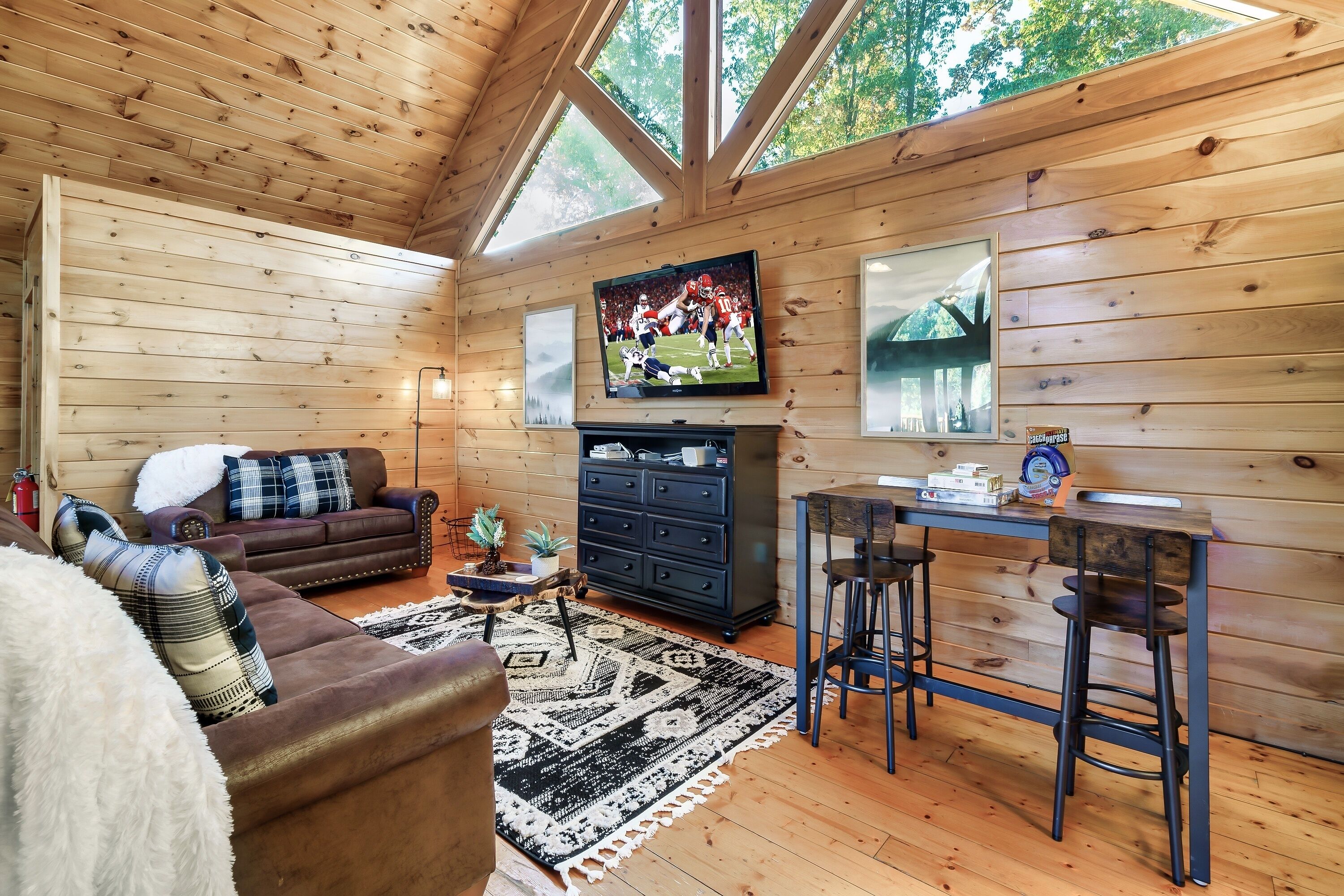 A cozy cabin interior with a wooden ceiling, a TV mounted above a fireplace, a sofa, a coffee table, and a bar area with stools.