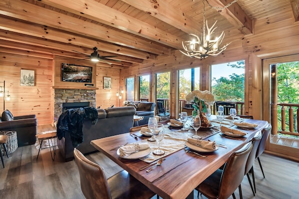 A wooden interior with a dining table set for six, a fireplace, and large windows offering a view of the outdoors.