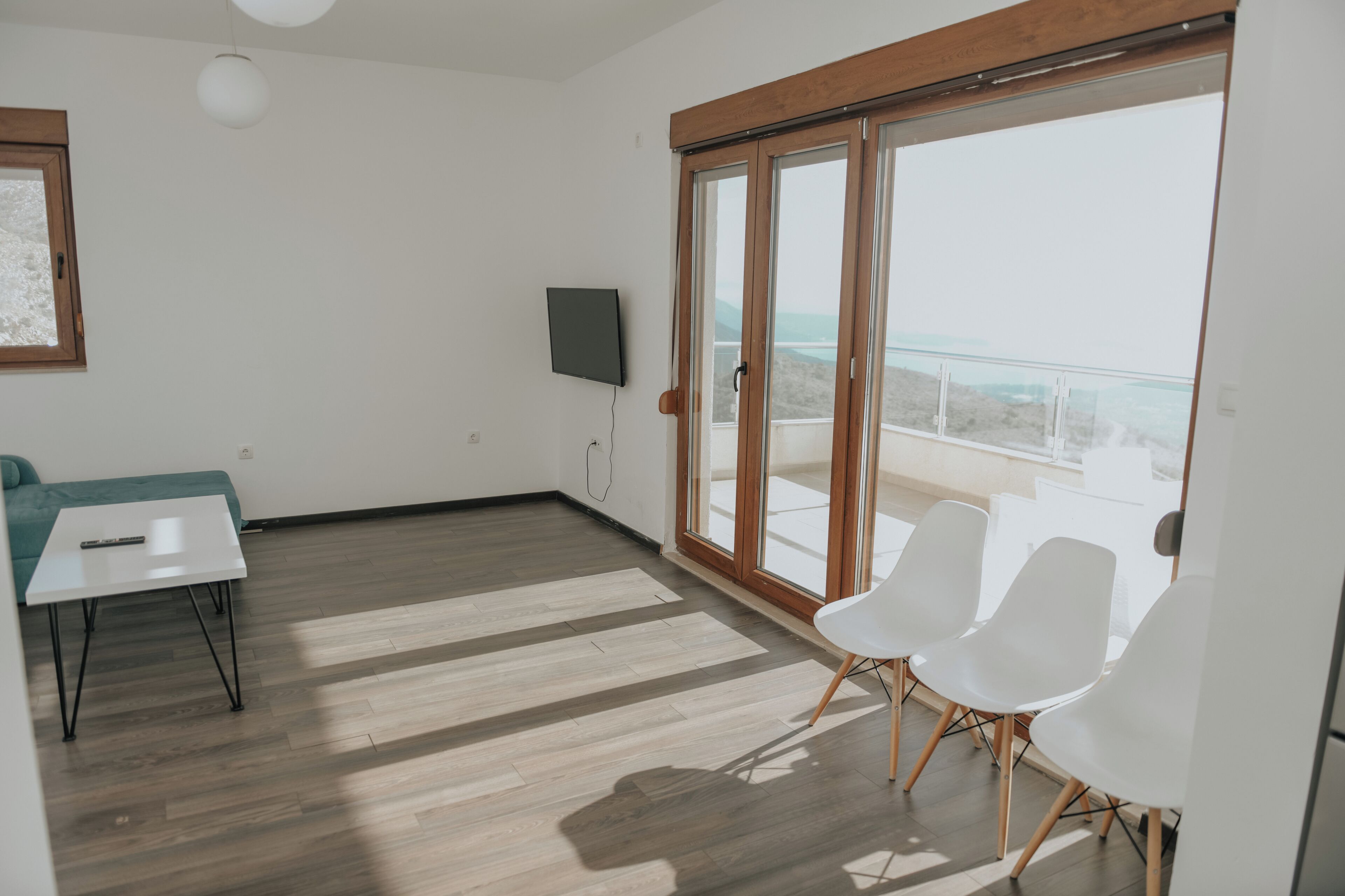 Quarto com grande porta de vidro deslizante que dá para uma varanda, televisão de tela plana na parede, mesa branca com controle remoto e duas cadeiras brancas.