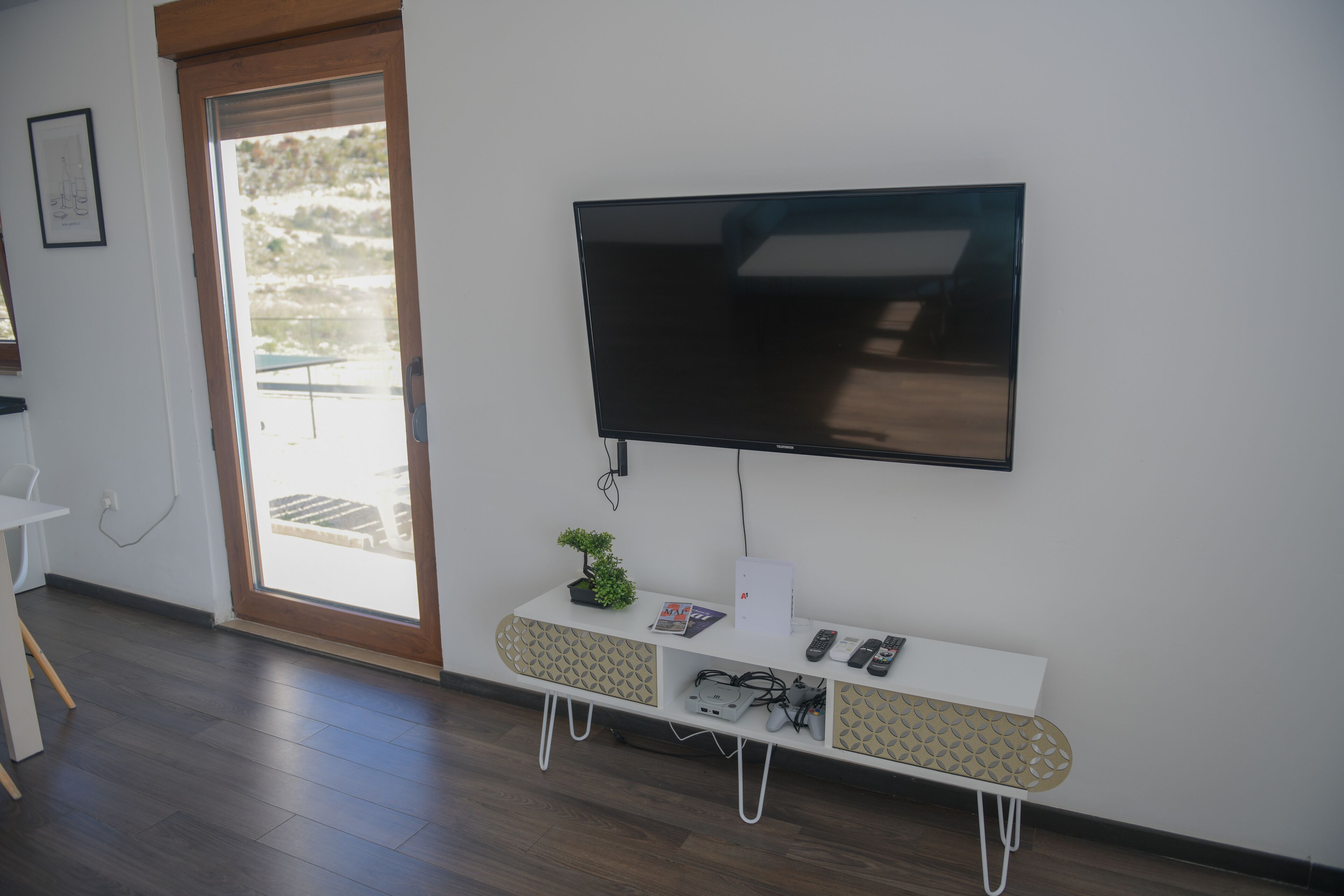 Un salon moderne avec un téléviseur à écran plat fixé au mur, une console blanche agrémentée d’une plante et une porte-fenêtre coulissante donnant sur un espace extérieur.