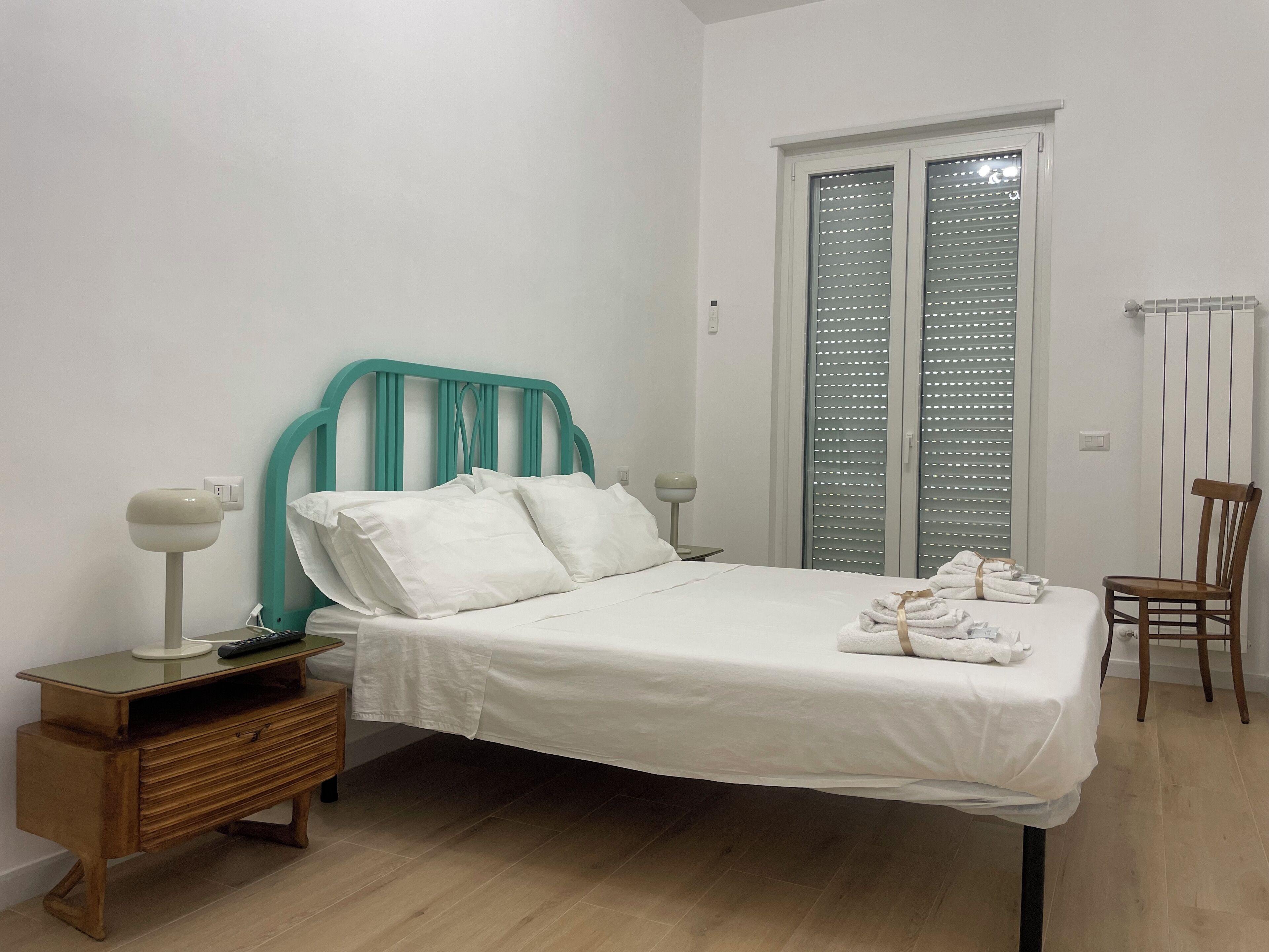 Una cama bien hecha con ropa blanca y almohadas, una mesita de noche de madera con una lámpara, una silla de madera y una ventana con persianas.