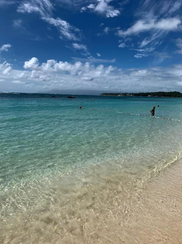 Una playa con aguas turquesas cristalinas, una orilla arenosa y una costa distante con edificios y vegetación.