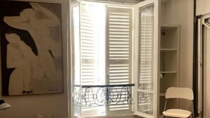 Et værelse med seng, radiator, et vindue med skodder, en stol og et billede på væggen.