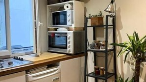 Et kompakt køkken med komfur, mikroovn og et lille køleskab. Der er et vindue, der giver naturligt lys, og en potteplante på en hylde.