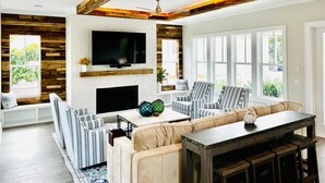 A modern living room with a fireplace, a large TV, striped sofas, and a wooden ceiling structure. - Toes in the Water (Rosemary Beach)