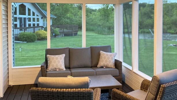 Eine überdachte Veranda mit einer Couch und Korbstühlen, großen Fenstern und Blick auf ein Haus und Garten.