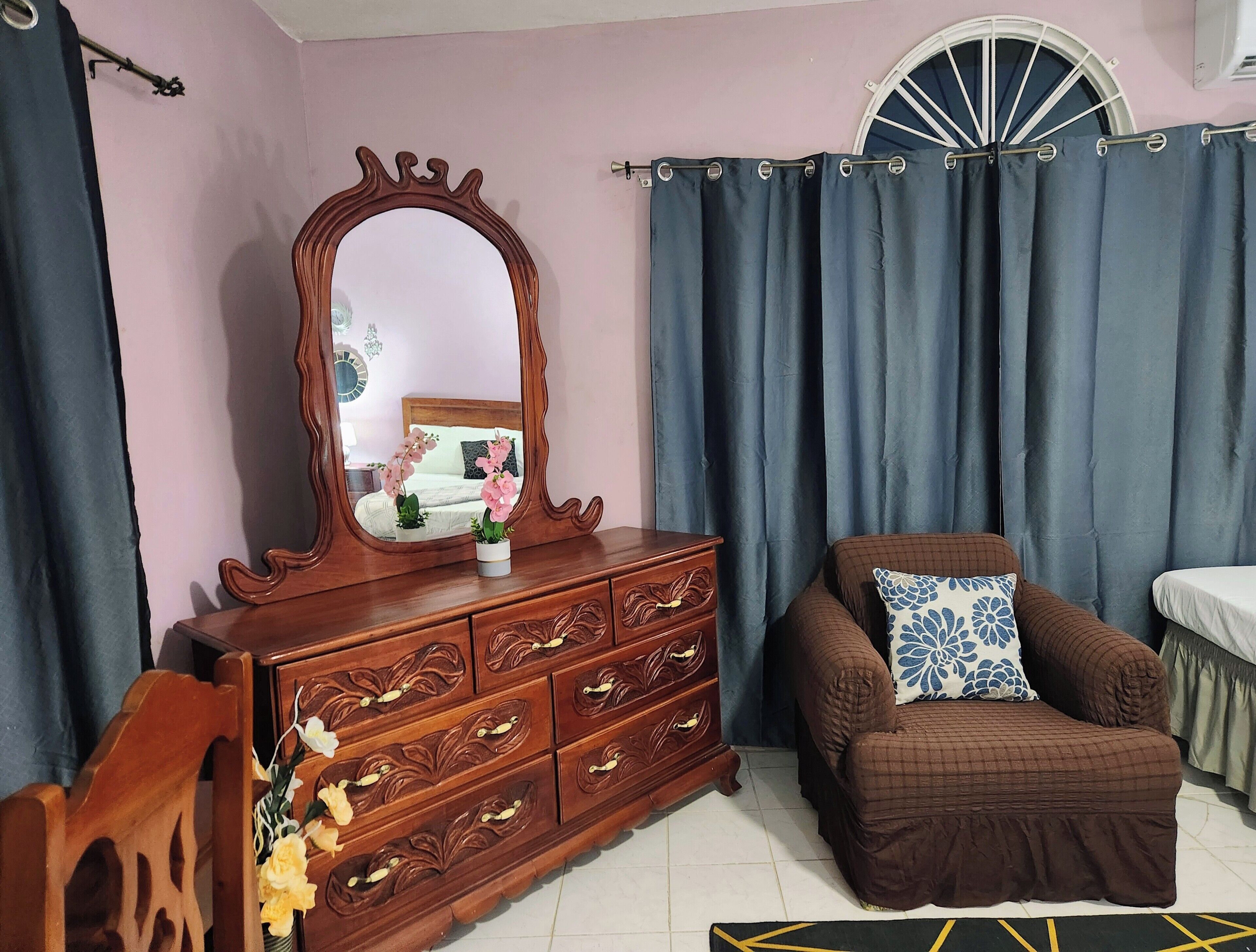 A bedroom with a wooden dresser, a mirror, a brown sofa with a patterned pillow, and a window with curtains.