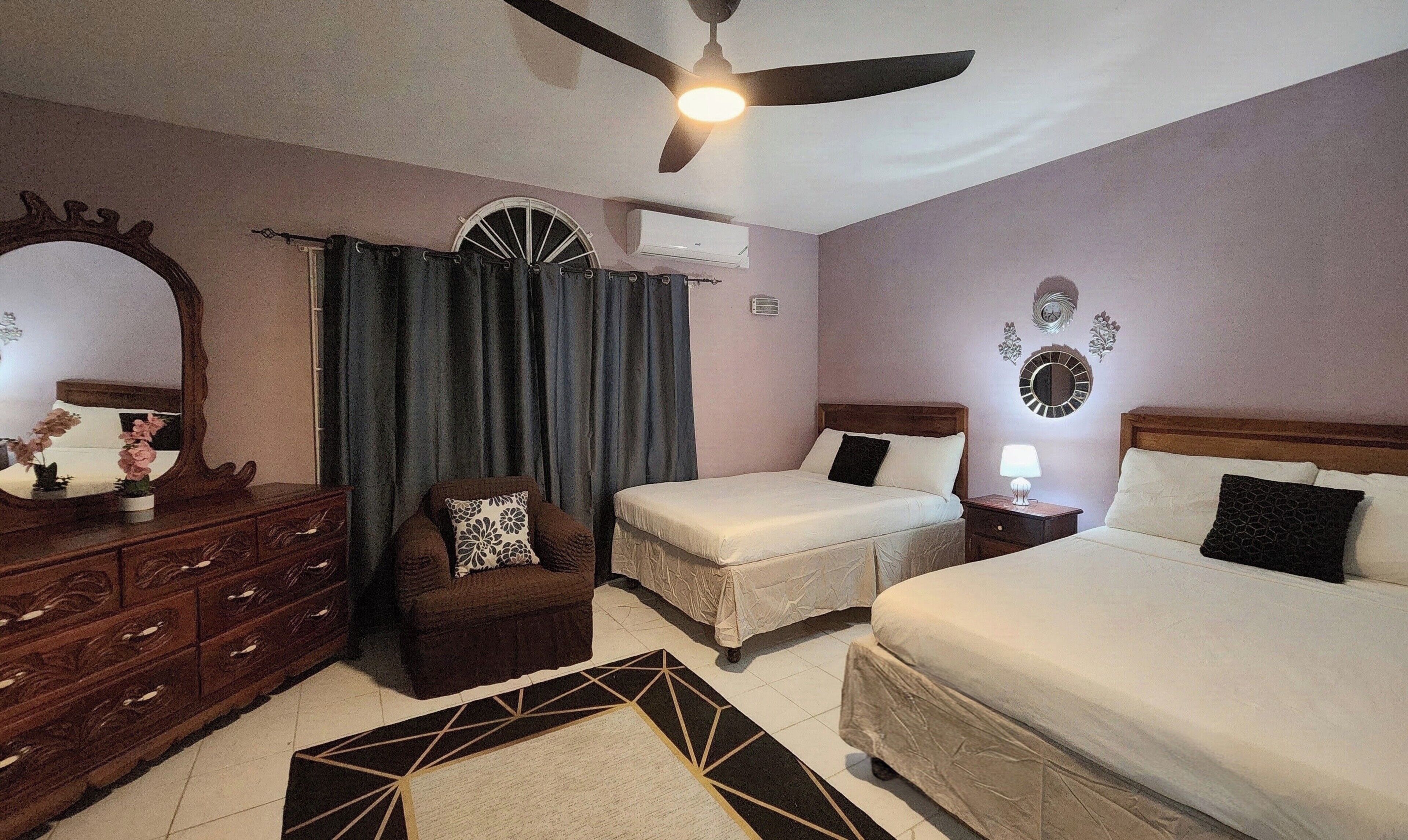 Habitación de hotel con dos camas, ventilador de techo, espejo, cómoda y ventana con cortinas.