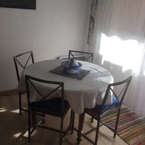 Una mesa redonda de comedor con mantel blanco, rodeada por cuatro sillas con estructura de madera y cojines oscuros. Un objeto decorativo azul está sobre la mesa.