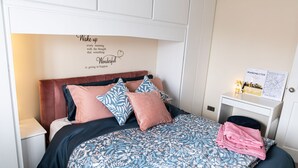 A neatly made bed with a floral-patterned bedspread and multiple pillows, a white nightstand with a lamp, and a wall with a motivational quote. - 3 Bed House - Sleeps 6 - Garden - Parking - Wifi (Manchester)
