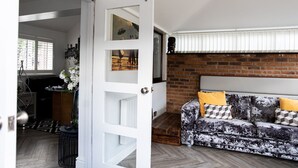 A room with a white door, a brick wall, a sofa with patterned cushions, and a window with blinds. - 3 Bed House - Sleeps 6 - Garden - Parking - Wifi (Manchester)