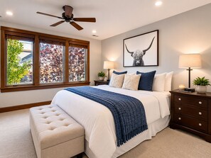 Une chambre à coucher avec un lit, un ventilateur de plafond, une fenêtre donnant sur des arbres, une table de chevet avec une lampe et un tableau encadré au mur.