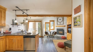 Een keuken met houten kastjes, een vaatwasser en hangende lampen. Er is een eethoek met een tafel en stoelen, en een open haard.