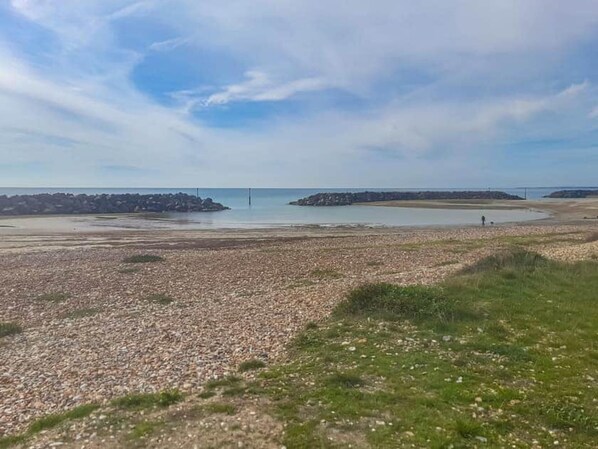 Un paysage côtier avec une plage de galets, des zones herbeuses et une vue lointaine sur la mer.
