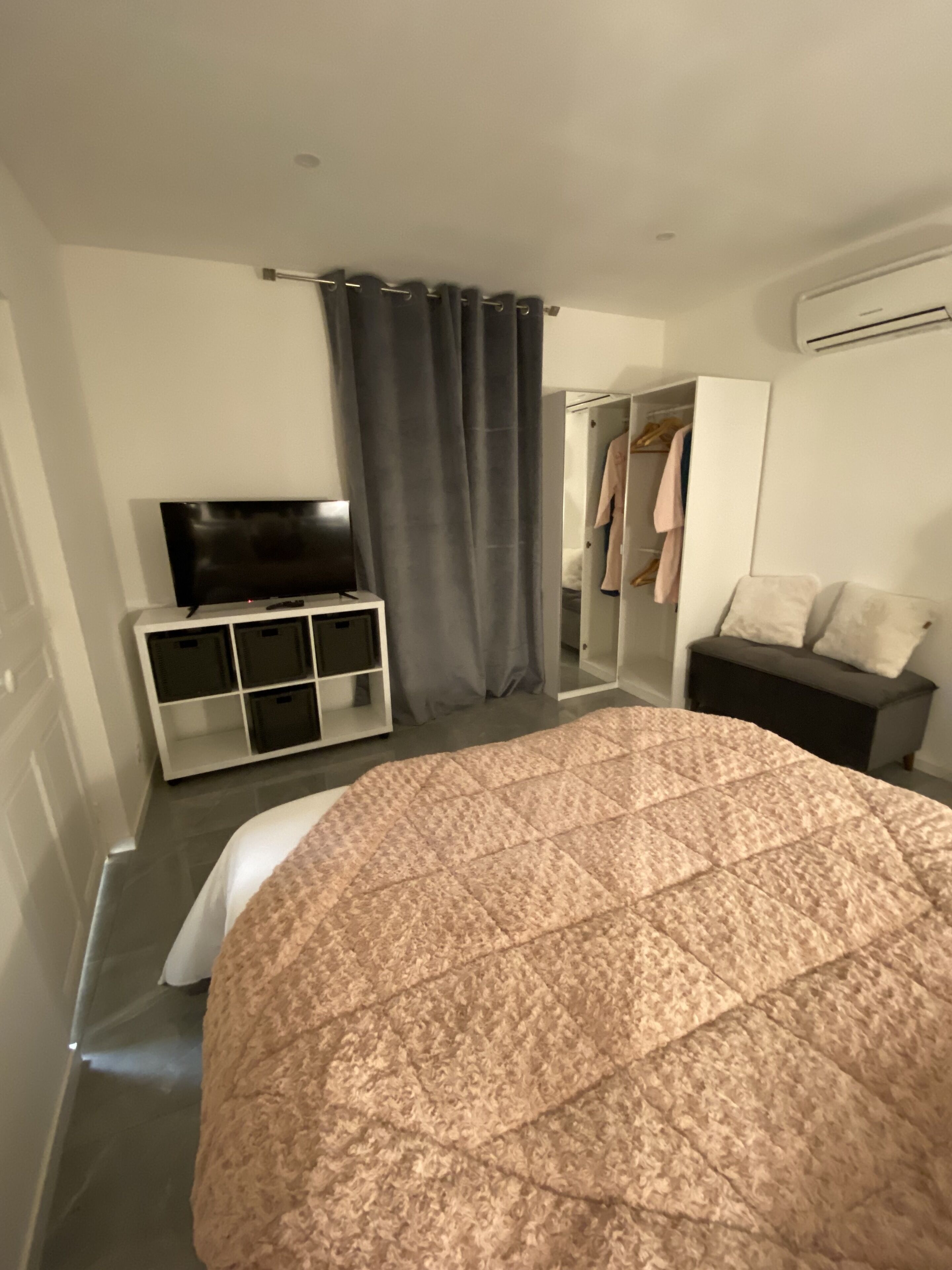 Una habitación de hotel moderna con una cama grande, un televisor sobre un soporte blanco, un armario y una zona de ducha.