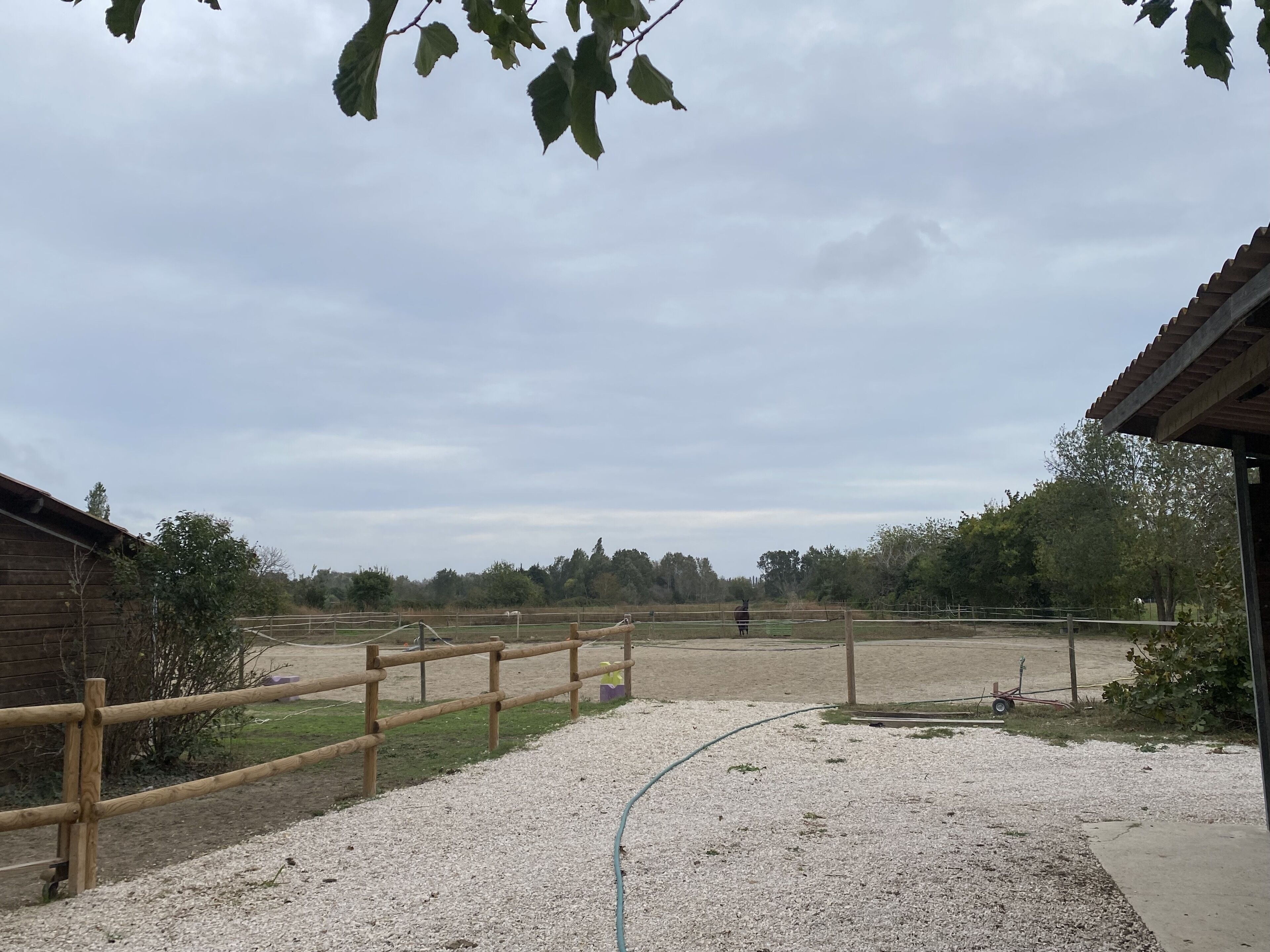 A stable with a wooden fence, a gravel path, and a horse in the background.