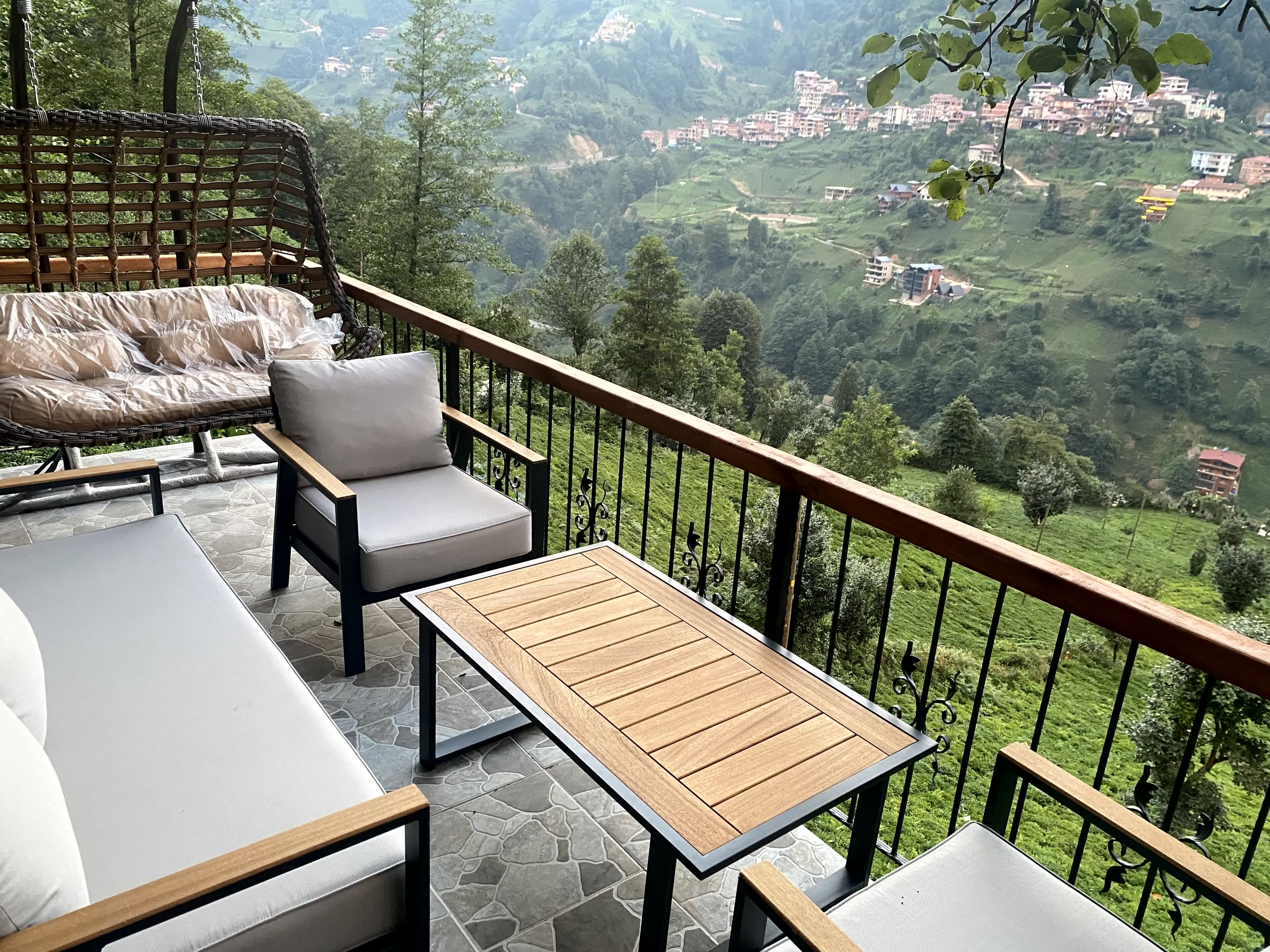 A balcony with a cushioned seating area, a wooden table, and a view of a hilly village.