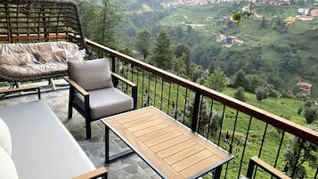 A balcony with a cushioned seating area, a wooden table, and a view of a hilly village.