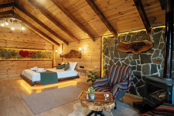 A cozy wooden cabin interior with a bed, a colorful armchair, a stone wall, and a fireplace. - Mezra Dogal Dag Evleri (Rize)