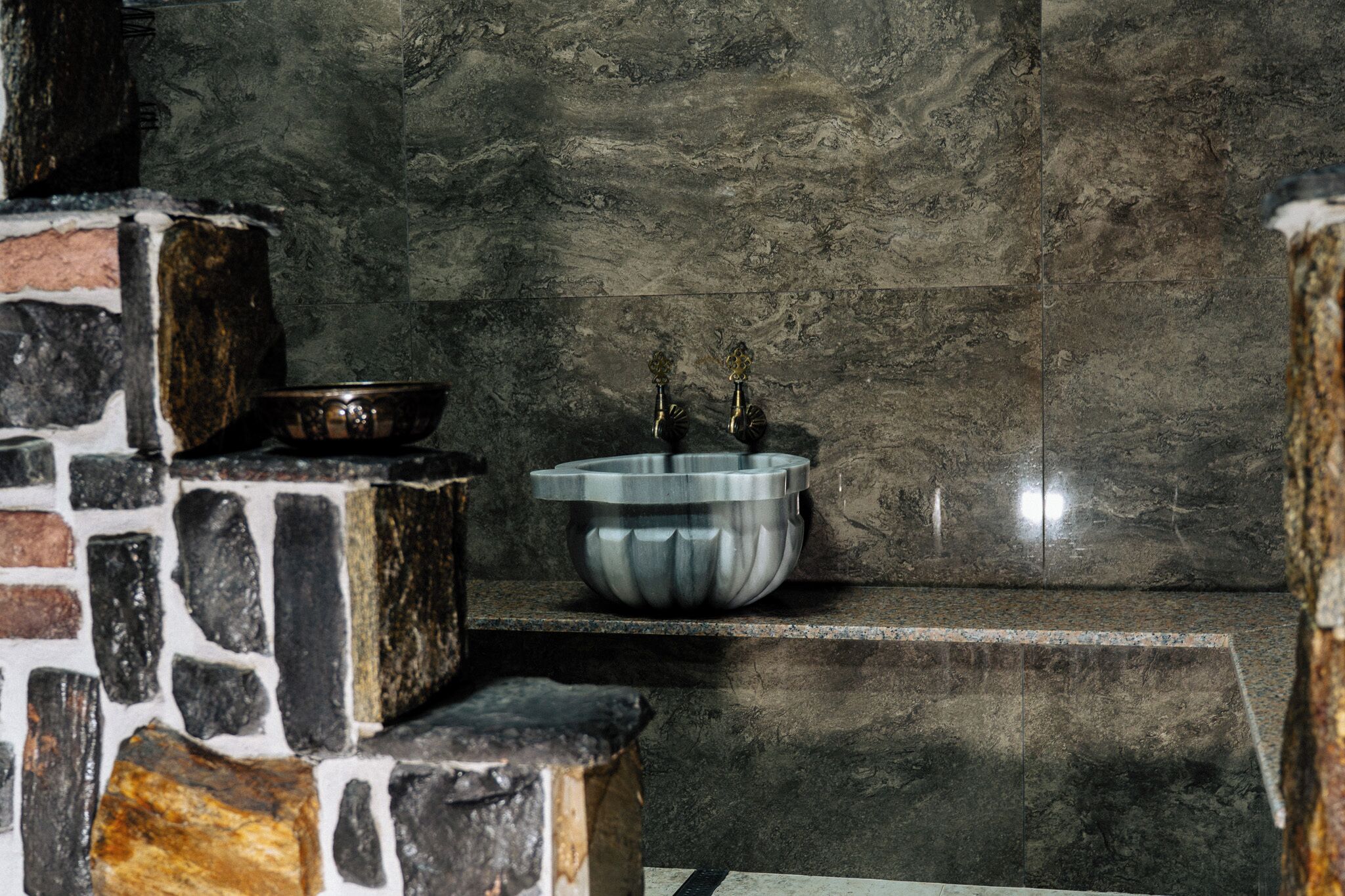A bathroom with a stone wall, a metal sink, and a bowl on a shelf.