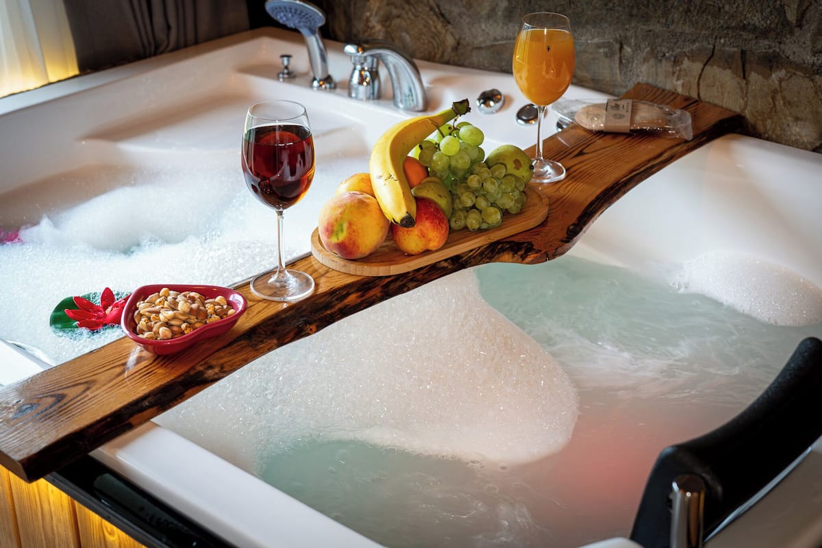 A bathtub with a wooden tray holding fruits and glasses of wine.