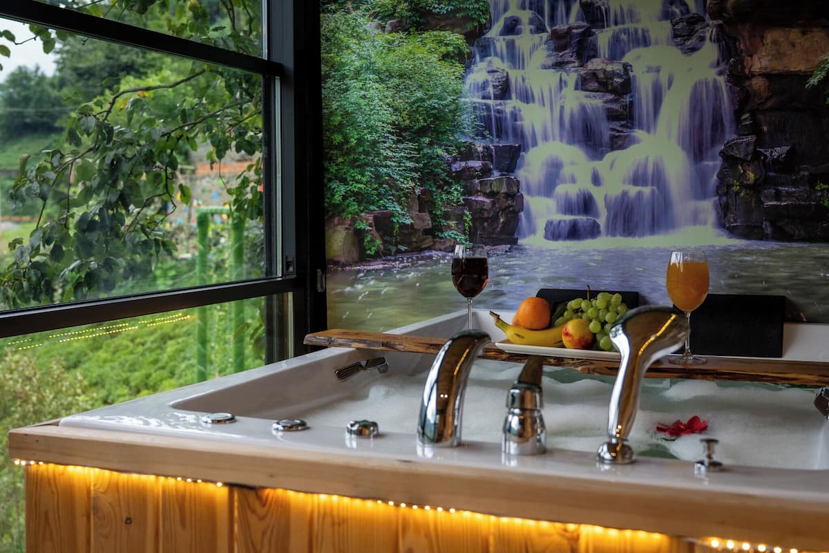 A bathroom with a sink, two faucets, and a window overlooking a waterfall.